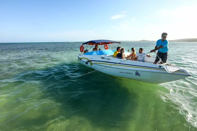 Um barco pequeno com algumas pessoas sendo guiado por um marinho sobre um mar esverdeado em Boipeba onde fica