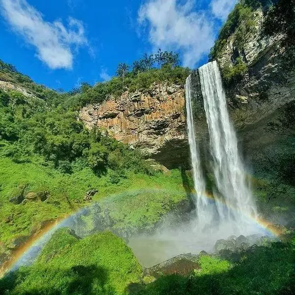 A cachoeira do Caracol, com sua queda d'água a frente de um paredão coberto de vegetação nativa, sob um céu azul e com um arco-íris em sua base. Uma das cachoeiras mais bonitas do Brasil.