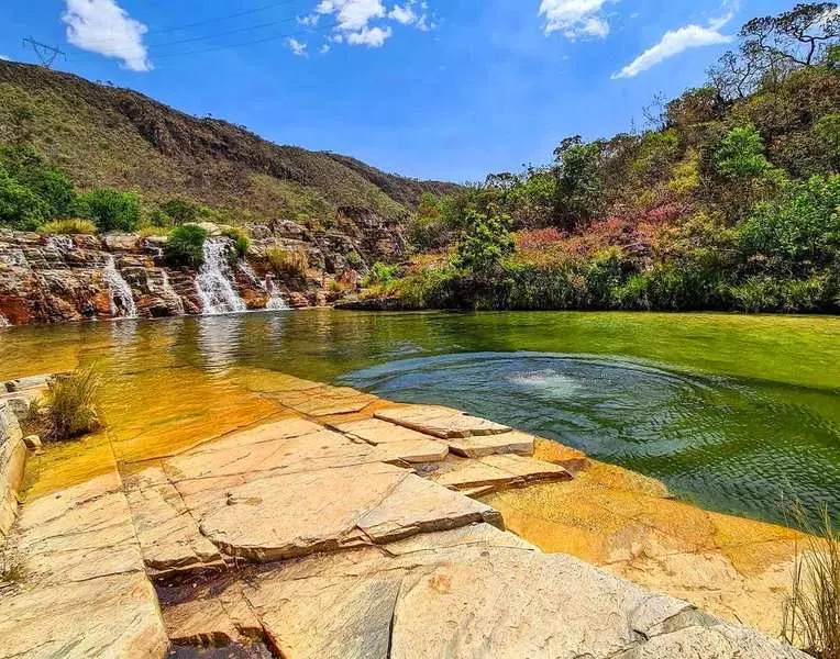 Enorme piscina natural com bordas naturais de rocha. Ao longe, a cobertura vegetal é um pequeno paredão de rochas com algumas quedas d'água de cachoeiras.
