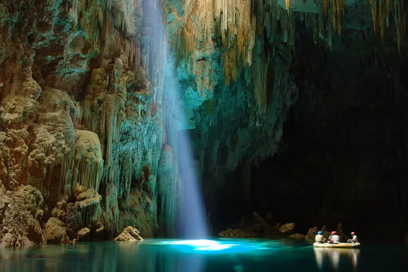 Caverna com feixe de luz natural iluminando rio subterrâneo ao lado de um balsa com pessoas. Bonito, em Paredão rochoso, com duas quedas d'água que desaguam em um lago na Chapada dos Veadeiros em lugares para viajar no brasil