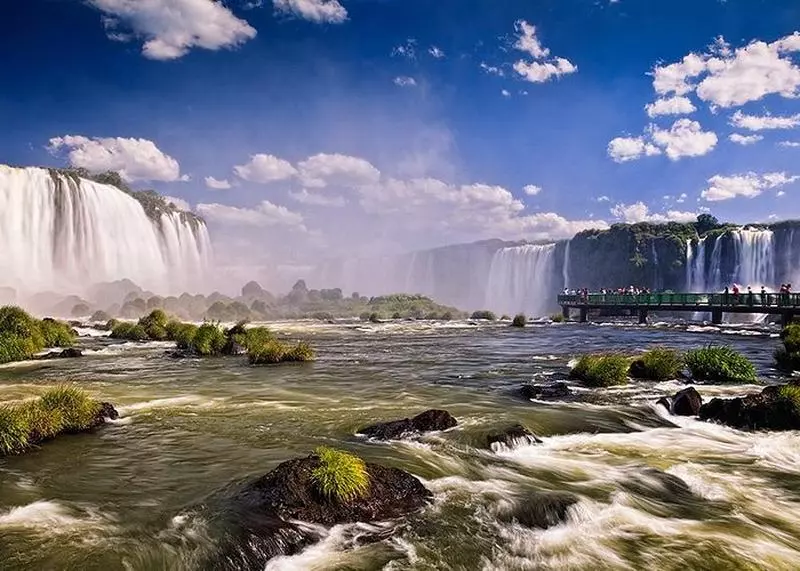 Panorâmica das Cataratas de Foz do Iguaçu, mostra as quedas de água a distância junto da ponto que turistas usam para ter acesso a elas. Um dos Paredão rochoso, com duas quedas d'água que desaguam em um lago na Chapada dos Veadeiros em lugares para viajar no brasil