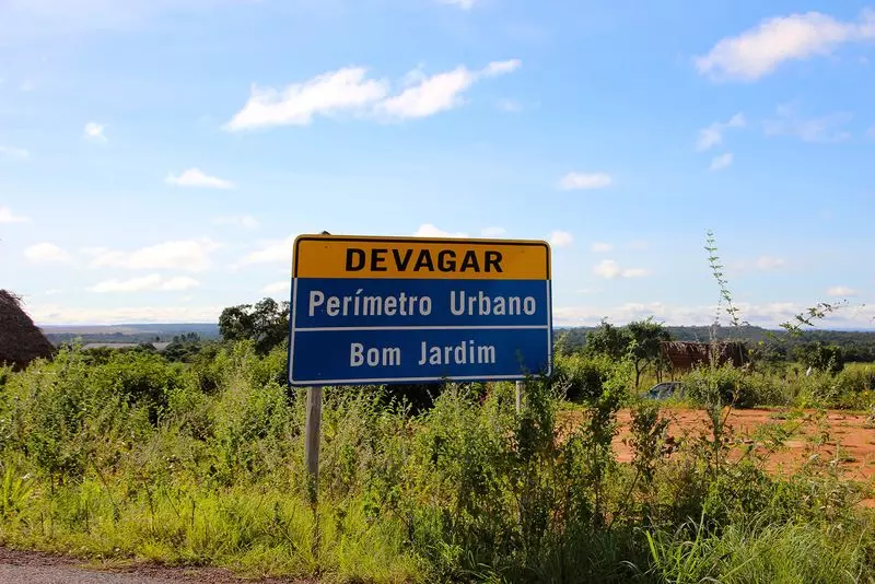 Nobres MT, placa notificando chegada ao perímetro urbano de Bom Jardim em meio a vegetação rasteira na lateral da rodovia.